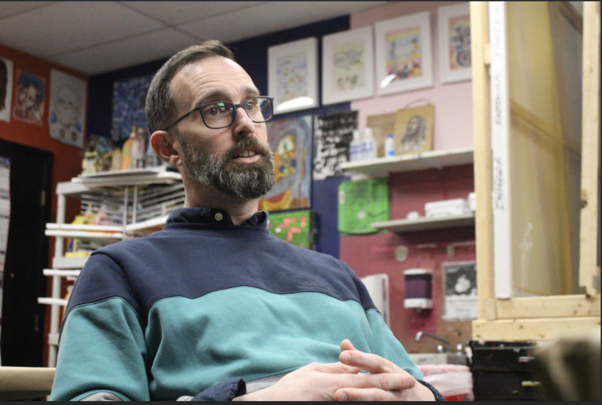 A man with glasses and a beard sits with hands clasped in a room filled with artwork and office supplies. He is wearing a blue and green sweater.